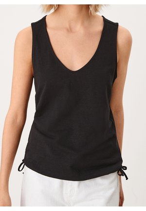 Ærmeløs sort V-hals top med snore i siderne båret af person med lys hud og hvide bukser mod ensfarvet baggrund.