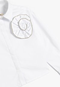 Camisa blanca con un destacado detalle de flor rizada blanca y ribete plateado en el pecho. Fabricada en tejido suave con un collar estructurado.