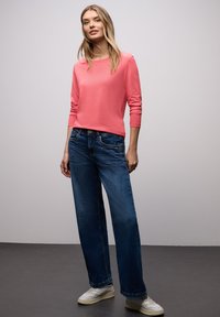 Koralowy top z długim rękawem zestawiony z niebieskimi dżinsami o szerokich nogawkach i białymi trampkami. Gładka tkanina, luźny krój i prosty design.