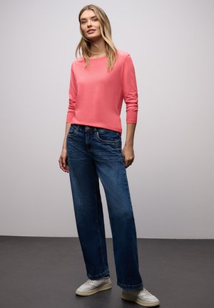 Korallenfarbenes Langarmshirt kombiniert mit blauen Jeans mit weitem Bein und weißen Turnschuhen. Glatter Stoff, lockere Passform und schlichtes Design.