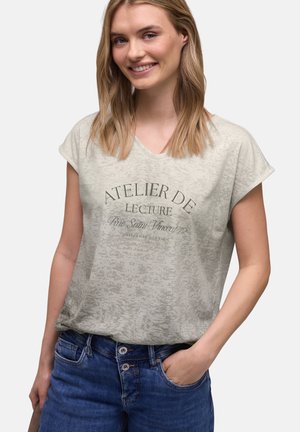 Smilende kvinde med skulderlangt blondt hår iført en beige t-shirt med print og blå jeans, stående med den ene hånd i lommen.