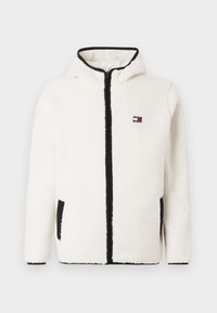 Valkoinen fleece-takki, jossa on huppu, mustat reunat ja vetoketjusuljenta. Takissa on sivutaskut ja pieni logo rinnassa. Pehmeä, teksturoitu materiaali.