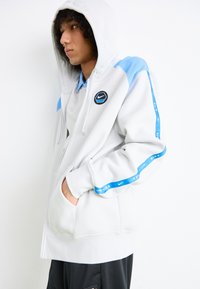 Sweatshirt com fecho da Nike em cinza claro com detalhes em azul, apresentando um logo redondo no peito, bolsos laterais e punhos e bainha canelados.