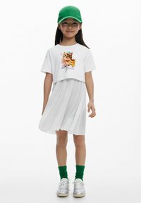 Fehér crop t-shirt macska grafikával, ami egy laza fehér pliszézett szoknyával van párosítva. Zöld sapka és zoknik; ezüst tornacipő egészíti ki a megjelenést.