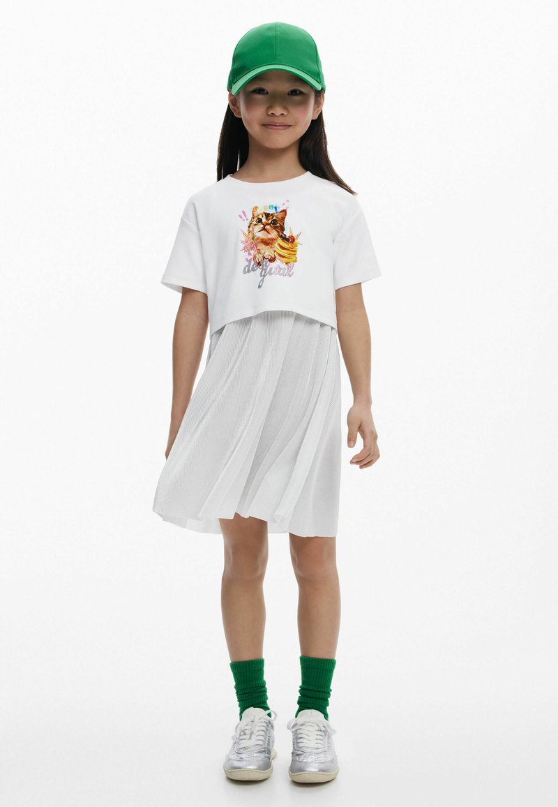 Fehér crop t-shirt macska grafikával, ami egy laza fehér pliszézett szoknyával van párosítva. Zöld sapka és zoknik; ezüst tornacipő egészíti ki a megjelenést.