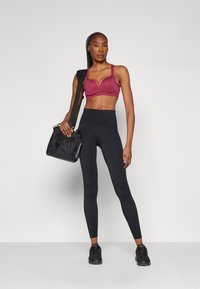 Burgunder sports-BH med V-formet utringning, svarte high-waisted leggings med perforerte detaljer, og en svart skulderveske.