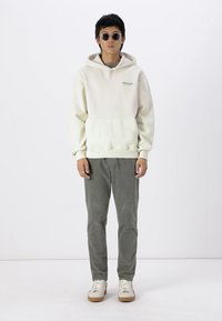 Cremfarbener Hoodie mit einer Vordertasche, kombiniert mit grauen Cordhosen und weißen Sneakern mit braunen Sohlen. Der Text auf dem Hoodie ist dezent.