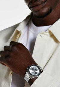 Zilveren chronograafhorloge met zwarte accenten, roestvrijstalen armband en een witte wijzerplaat. Draagt aan een donkerharige hand, naast een lichte jas.