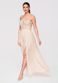 Femme portant une robe bustier sans manches beige clair ornée de sequins, avec une fente haute à la cuisse, des talons ouverts assortis et un bracelet.