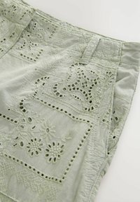 Tissu vert clair avec des motifs brodés complexes et des détails découpés, présentant une coupe décontractée et des coutures visibles le long des bords.