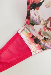 Bikini zgornji del s cvetličnim vzorcem v beli in rdeči barvni shemi. Ima mehko tkanino, drzne roza poudarke in vidne detajle šivanja.