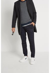 Donker wollen overjas, gestreepte marineblauwe trui, geruite marineblauwe broek en witte sneakers. Accessoires zijn zwarte zonnebrillen die in de hand worden vastgehouden.
