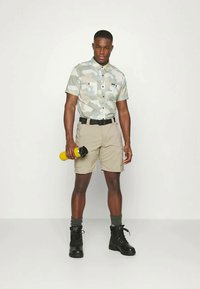 Camisa de manga curta em camuflagem cinza claro, calções bege, cinto preto, botas de combate pretas, segurando uma garrafa de água amarela, de pé sobre um fundo simples.