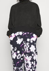 Osoba oblečená v černé texturované zkrácené mikině a fialovo-bílých květinových kalhotách Adidas, má ruce v kapsách.