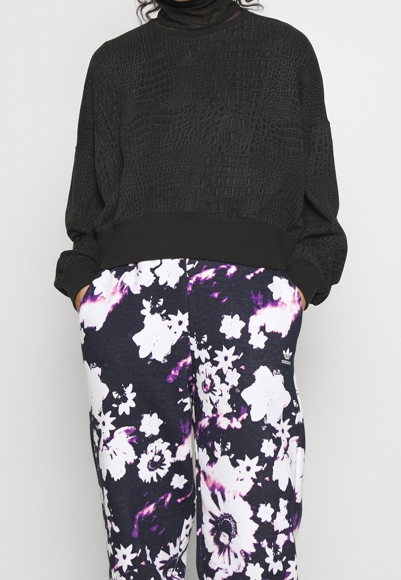 Osoba oblečená v černé texturované zkrácené mikině a fialovo-bílých květinových kalhotách Adidas, má ruce v kapsách.