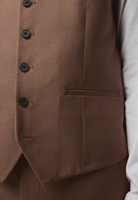 Gilet en brun texturé présentant cinq boutons noirs et une poche latérale. Le tissu a un motif subtil, offrant une apparence sur mesure.
