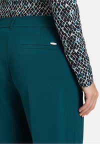 Teal hoge taillebroek met een gladde textuur, voorzien van een achterzak en een zilveren merklabel. Gepaard met een gestreepte zwart en teal top.