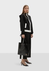 Crna kapuljača s bijelim detaljima, crna torba, crne cipele s oštrim vrhom, nosi žena koja stoji protiv svijetle pozadine.