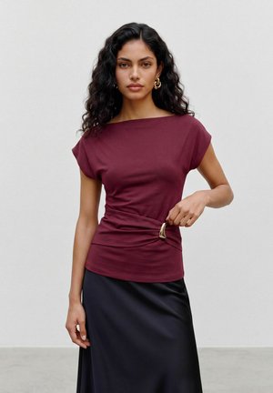 Femme aux longs cheveux bouclés portant un haut bordeaux à manches courtes avec une boucle dorée et une jupe noire, debout devant un fond uni.
