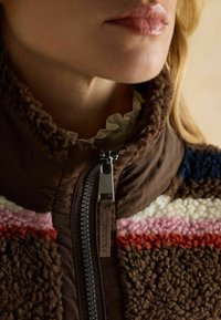 Braune Fleecejacke mit hohem Kragen, versehen mit horizontalen Streifen in Rot, Pink, Weiß und Blau. Vorderer Reißverschluss mit einem Detail aus Stoffetikett.