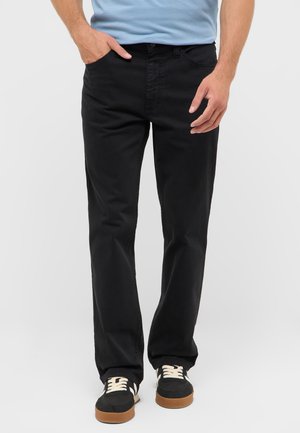Mann trägt schwarze Straight-Fit-Jeans, hellblaues Hemd und schwarze Sneakers mit weißen Schnürsenkeln und steht vor einem schlichten Hintergrund.
