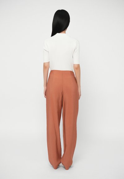 Λευκή ριγέ μπλούζα κοντομάνικη συνδυασμένη με ψηλόμεσα, φαρδιά παντελόνια σε χρώμα οξειδωμένου πορτοκαλιού, αναδεικνύοντας μια λεία υφή και καθαρές γραμμές.