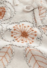 Vyšívaná látka s oranžovými květinovými a listovými vzory na světle béžovém pozadí, s černými stehy obrysujícími vzory.