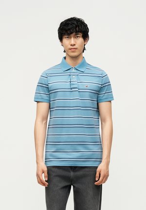 Giovane uomo che indossa una polo blu a maniche corte con righe bianche e blu scuro, in piedi davanti a uno sfondo chiaro uniforme.