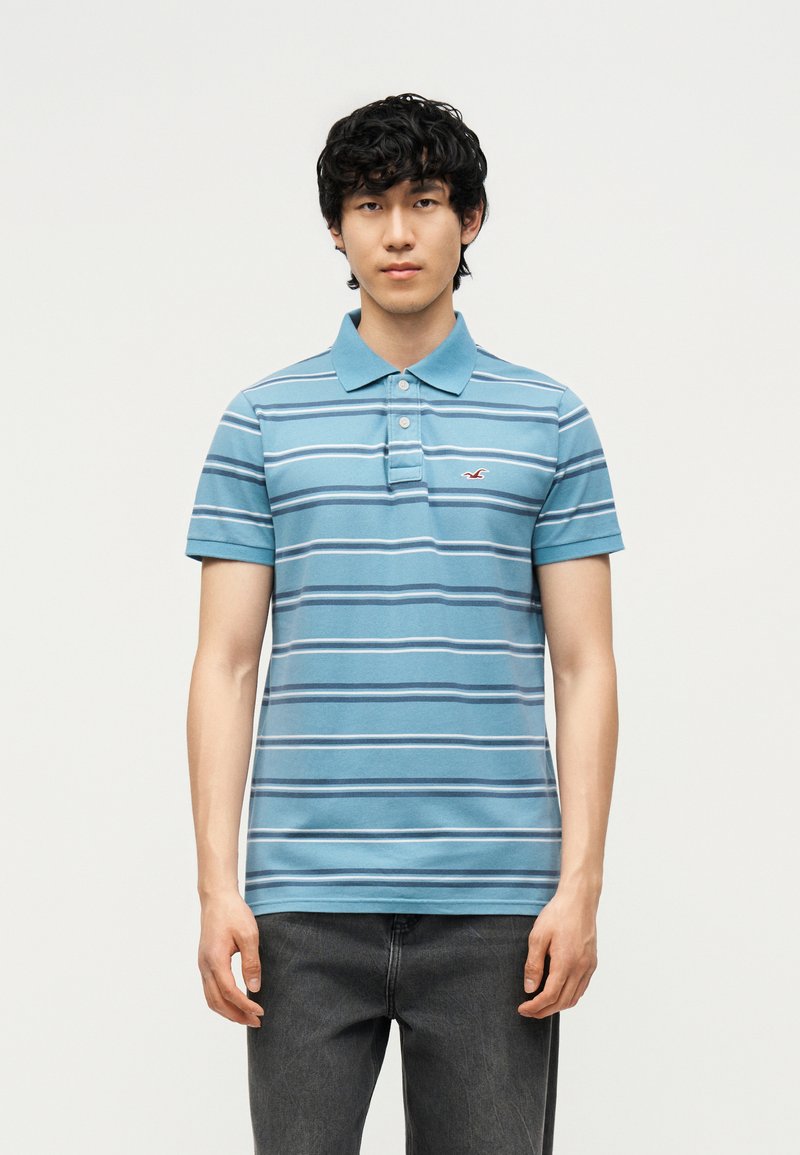 Jeune homme portant un polo bleu à manches courtes avec des rayures blanches et bleu foncé, debout devant un fond clair uni.