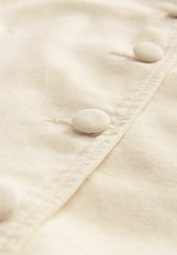 Un tissu crémeux texturé présente une patte de boutonnage avec des boutons ronds recouverts de tissu, mettant en avant des coutures subtiles et une surface douce et lisse.