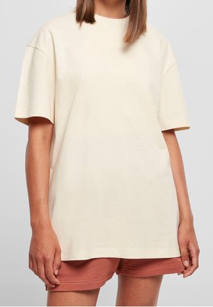 Kvinde iført en løs, cremefarvet t-shirt med korte ærmer og rustfarvede shorts mod en ensfarvet lysegrå baggrund.