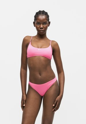 Γυναίκα όρθια, φοράει ροζ μπικίνι με λεπτές τιράντες, σε απλό λευκό φόντο.