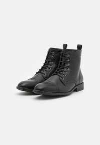 Botas de cuero negro hasta el tobillo con punta redondeada, parte frontal con cordones, dos lengüetas de agarre y detalles de costura. Suela plana de goma para durabilidad y comodidad.