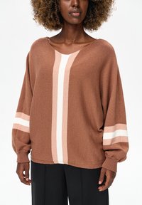 Vrouw draagt een losse bruine gebreide trui met roze en witte verticale en horizontale strepen, gecombineerd met een zwarte broek, tegen een witte achtergrond.