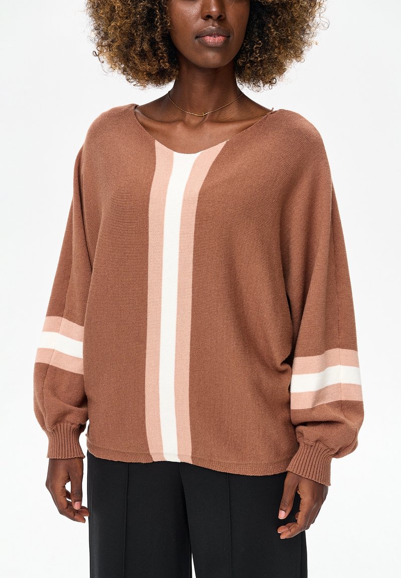 Vrouw draagt een losse bruine gebreide trui met roze en witte verticale en horizontale strepen, gecombineerd met een zwarte broek, tegen een witte achtergrond.