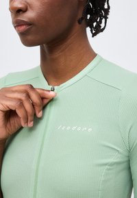 Άτομο με σκουρόχρωμο δέρμα και κοντά dreadlocks κλείνει το φερμουάρ ενός ανοιχτό πράσινου υφασμάτινου ποδηλατικού μπλουζιού με το λογότυπο "Isadore" στο στήθος.