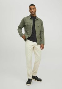 Giacca verde con bottoni, dotata di due tasche sul petto, indossata sopra una camicia nera. Pantaloni color crema e scarpe nere completano l'outfit.