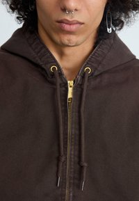 Felpa zip-up marrone realizzata in tessuto testurizzato, con zip dorata, lacci e cappuccio. Include un orecchino con spilla di sicurezza argentata.