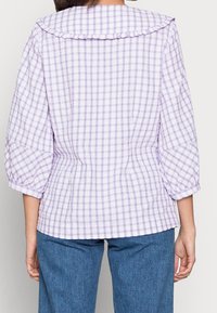 Lilla ternet bluse med flæsekrave, 3/4 puffede ærmer og figursyet talje, parret med blå denimbukser. Tekstureret bomuldsstof.