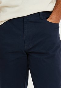 Dunkelblaues Baumwollhose mit glatter Oberfläche, Fronttaschen und sichtbaren Nahtdetails für einen maßgeschneiderten Look.
