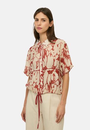 Mujer que lleva una camisa abotonada de manga corta con cuello y cintura de cordón, con un patrón abstracto beige y rojo, combinada con pantalones de color claro.