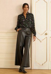 Melna ādas plaša jakas bikses ar augstu vidukli, kombinētas ar tumši zaļu rakstainu blūzi, kurai ir rozā akcenti. Modelis stāv uz gaiša fona.