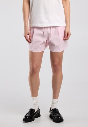 Personne portant un short à cordon de serrage rose clair, des chaussettes blanches montantes, des mocassins à plateforme noirs et un t-shirt blanc, debout devant un fond uni.