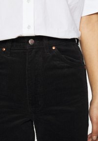 Černé kordové kalhoty s vertikálním žebrováním, s měděnými kovovými prvky a zapínáním na knoflík v pase. Klasický design s pěti kapsami.