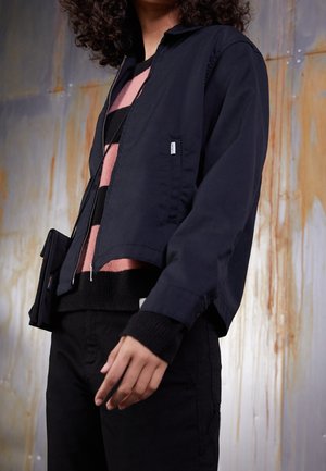 Person, die eine schwarze Jacke über einem pink-schwarz gestreiften Pullover, schwarze Hosen und eine kleine schwarze Umhängetasche trägt, vor einer verwitterten Wand.