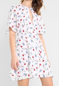 Vit kortärmad klänning med röd och blå blommig print, nyckelhålshalsringning och lagerad asymmetrisk nederdel, bärs av en person.