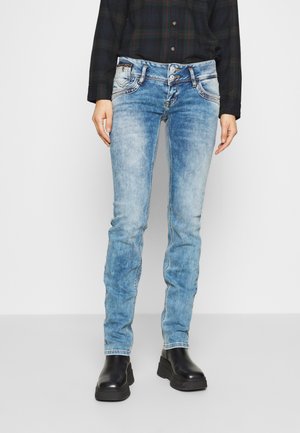 Personne portant un jean skinny bleu délavé, des bottes noires épaisses et une chemise sombre à manches longues avec boutons, debout devant un fond uni.