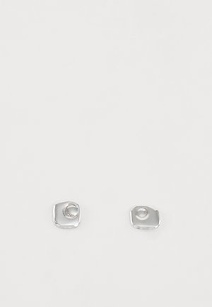 Deux petites boucles d'oreilles clous argentées en forme de carré avec des bords arrondis irréguliers sur un fond blanc uni.