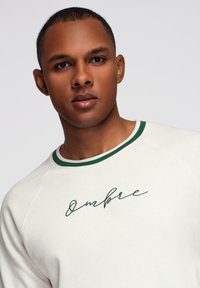 Joven con cabello corto que lleva un sudadera blanca con ribetes verdes y el texto "Ombre" bordado en el pecho, contra un fondo liso.