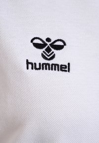 Witte stof met een zwart geborduurd logo met een bijontwerp en het woord "hummel" onder het logo, met een gestructureerd oppervlak.